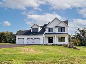 A two-story modern farmhouse-style home with white siding, black windows, and a three-car garage. The home sits on a spacious lot with a newly paved driveway, surrounded by green lawn and mature trees in the background.