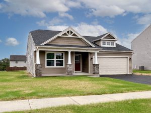 Single-story home with tan siding, stone accents on the columns, a front porch, and a two-car garage. Elmcrest model home.