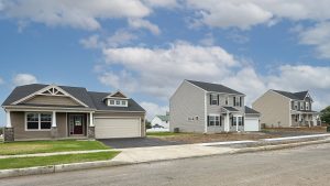 Street view showing three newly built homes, one ranch-style and two two-story models, in a row on a partially finished street. Model home grand opening this weekend. 