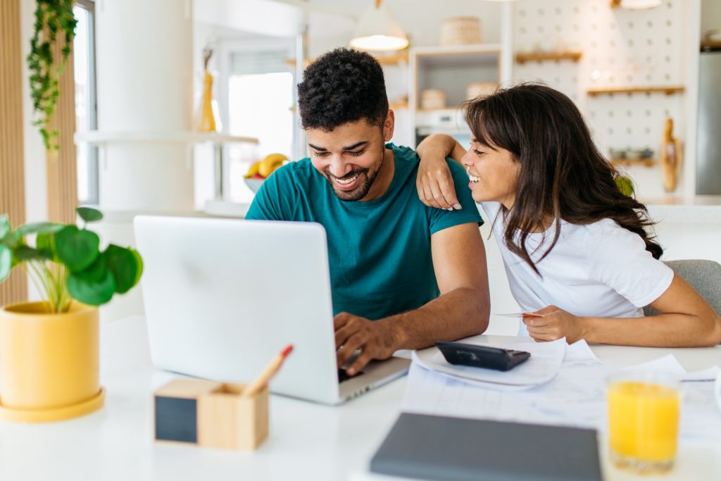 Tips to Save for Your Down Payment. A young responsible African American couple sits at home and calculating monthly income. The man is looking at the laptop while the woman explaining to him how to save some money.