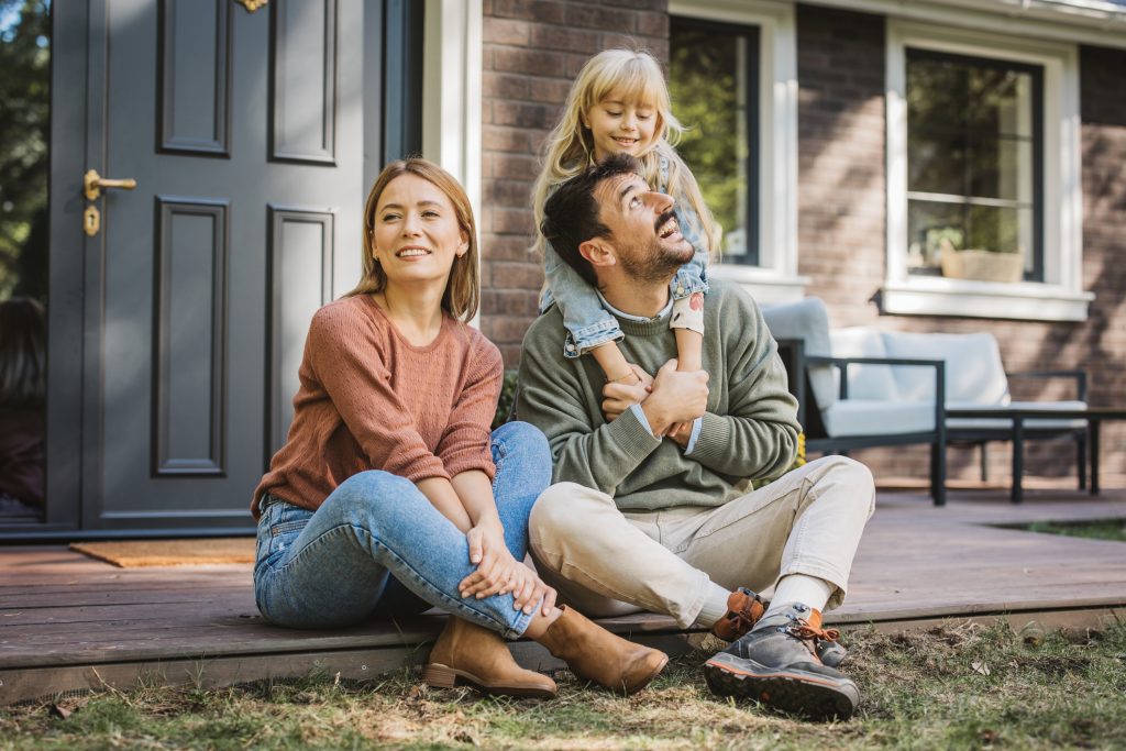 Tips to Save for Your Down Payment. Young family with little girl having fun in front of there house. Porch is decorated with flowers.